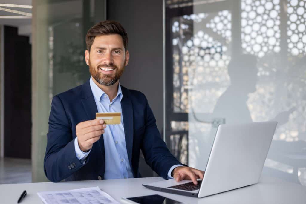 Portrait of smiling young man, office worker sitting at desk in office holding and showing credit card to camera